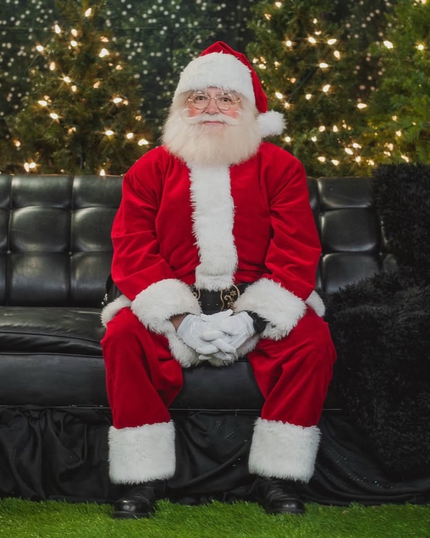 Santa seated on the black leather sofa in front of lit Christmas trees